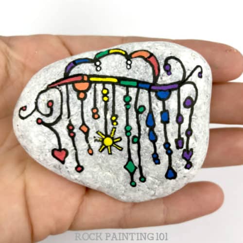 We are loving this rainbow zendangle painted rock! Let the colors of the rainbow dangle to create this beautiful and fun rock painting idea. #zendangle #rainbow #dangles #howtopaintrocks #howtozendangle #rainbowrocks #rockpainting #stonepainting #rockpainting101