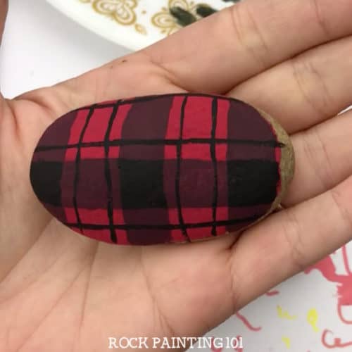 Learn how to paint plaid patterns with these step by step instructions and video tutorial. We used it to paint a fun Christmas rock, but you can use it for any project you have! #plaid #howtopaintplaid #flannel #red #christmasflannel #howtopaintrocks #christmasrockpaintingidea #tutorial #stonepainting #stockingstuffers #holidayrockhunting #giftideas #rockpainting101